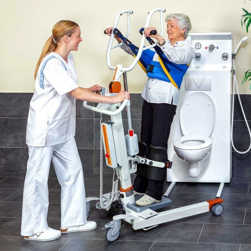 Podnośnik pionizujący NORA Alu podczas transferu pacjentki w łazience z toaletą.